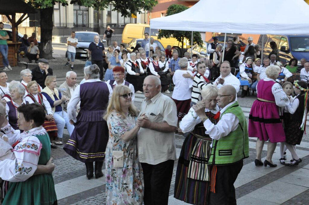 Duża grupa ludzi tańczy na rynku. W tle kilka drzew, samochody i elewacje budynków.