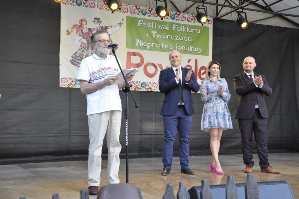 Kilka osób w oficjalnych strojach stoi na scenie plenerowej. Mężczyzna z siwą brodą trzyma podkładkę sceniczną i stoi przy mikrofonie na statywie, pozostali w dalszym planie klaszczą. Za nimi wisi baner promujący wydarzenie.