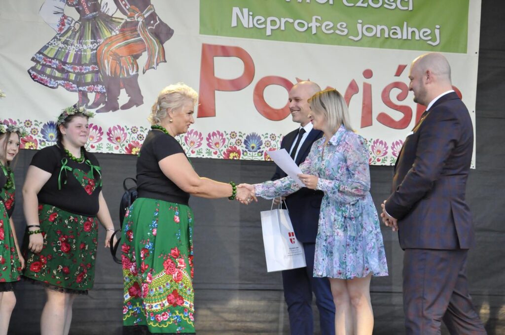 Kobieta w błękitnej kwiatowej sukience podaje dłoń kobiecie w ludowym stroju. Wokół więcej stojących uśmiechających się ludzi. W tle fragment baneru promującego wydarzenie.