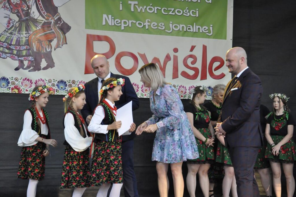 Kobieta w błękitnej kwiatowej sukience gratuluje dziewczynce w ludowym stroju. Wokół więcej stojących uśmiechających się ludzi. W tle fragment baneru promującego wydarzenie.