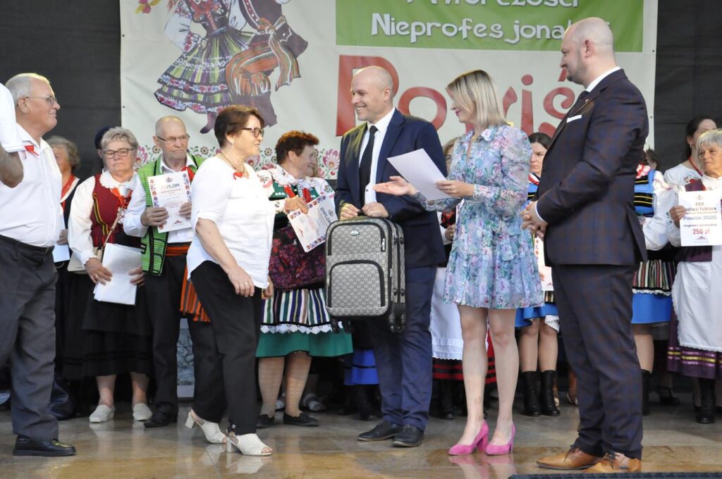 Mężczyzna w granatowym trzyma małą podróżną walizkę. Kobiet w błękitnej sukience trzyma dyplom.Wokół więcej stojących uśmiechających się ludzi. Niektórzy w strojach ludowych. W tle fragment baneru promującego wydarzenie.