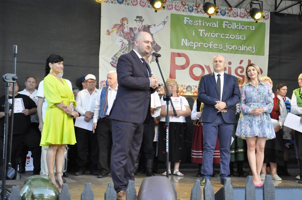 Łysy mężczyzna w granatowym garniturze stoi przy mikrofonie na statywie. Wokół stoi liczna grupa ludzi, niektórzy w ludowych strojach. W tle fragment baneru promującego wydarzenie.