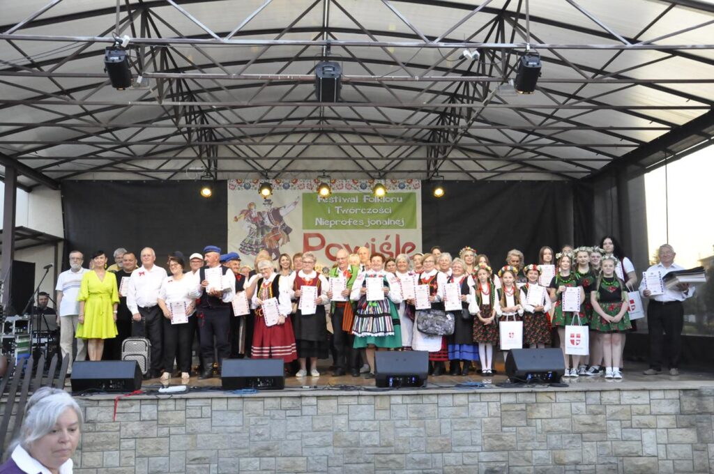 Liczna grupa dorosłych, młodzieży, dzieci i dorosłych pozuje do wspólnego zdjęcia na cenie plenerowej. Większość osób w strojach ludowych. Niektórzy trzymają dyplomy, inni statuetki. Tło czarno-granatowe. Nad nimi baner z nazwą wydarzenia.