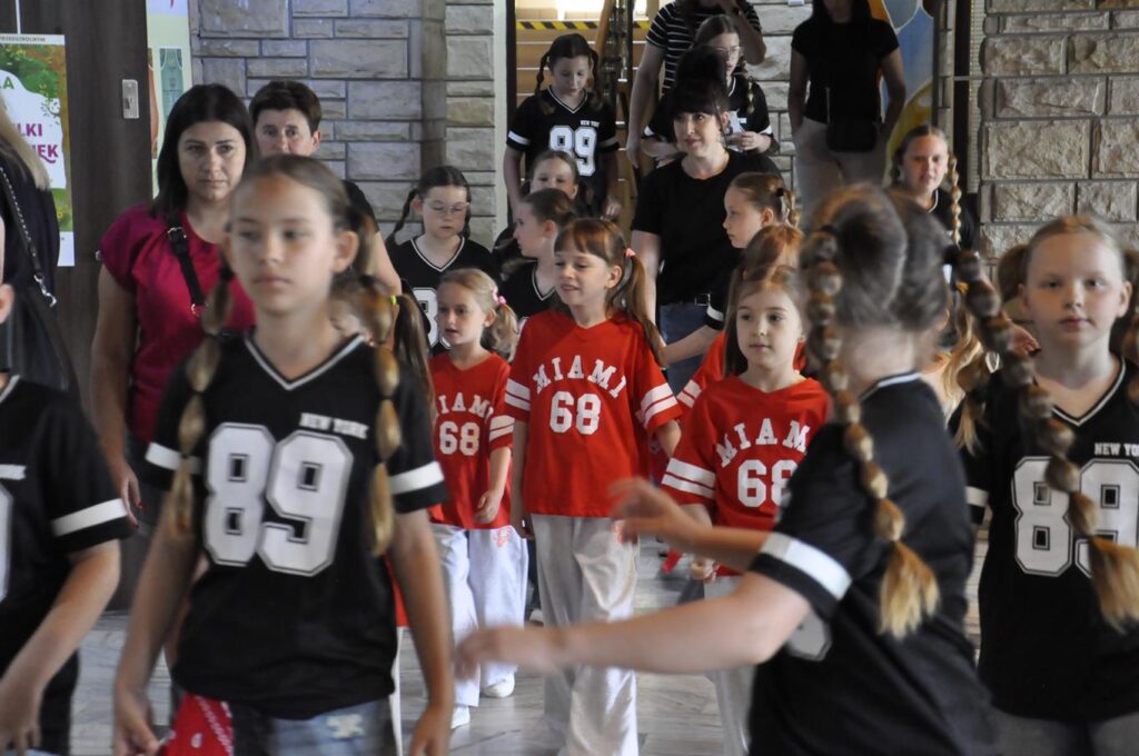Grupa młodzieży w czarnych i czerwonych strojach z numerami stłoczona w holu głównym budynku.