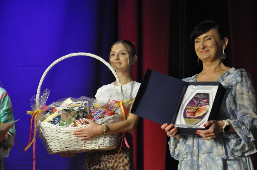 Kobieta w szarej, falbaniastej sukience trzyma etui ze statuetką Grand Prix. Druga kobieta w białej koszulce trzyma kosz ze słodyczami. Obie się uśmiechają. Tło bordowo-fioletowe.
