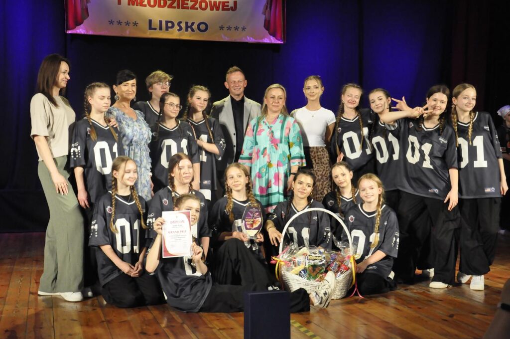 Grupa młodzieży w czarnych strojach z numerami pozuje do wspólnego zdjęcia z kilkoma dorosłymi. Jedna osoba wznosi dyplom. Przed nimi sto kosz ze słodyczami. Nad nimi fragment baneru z nazwą wydarzenia. Tło czarno-granatowe.