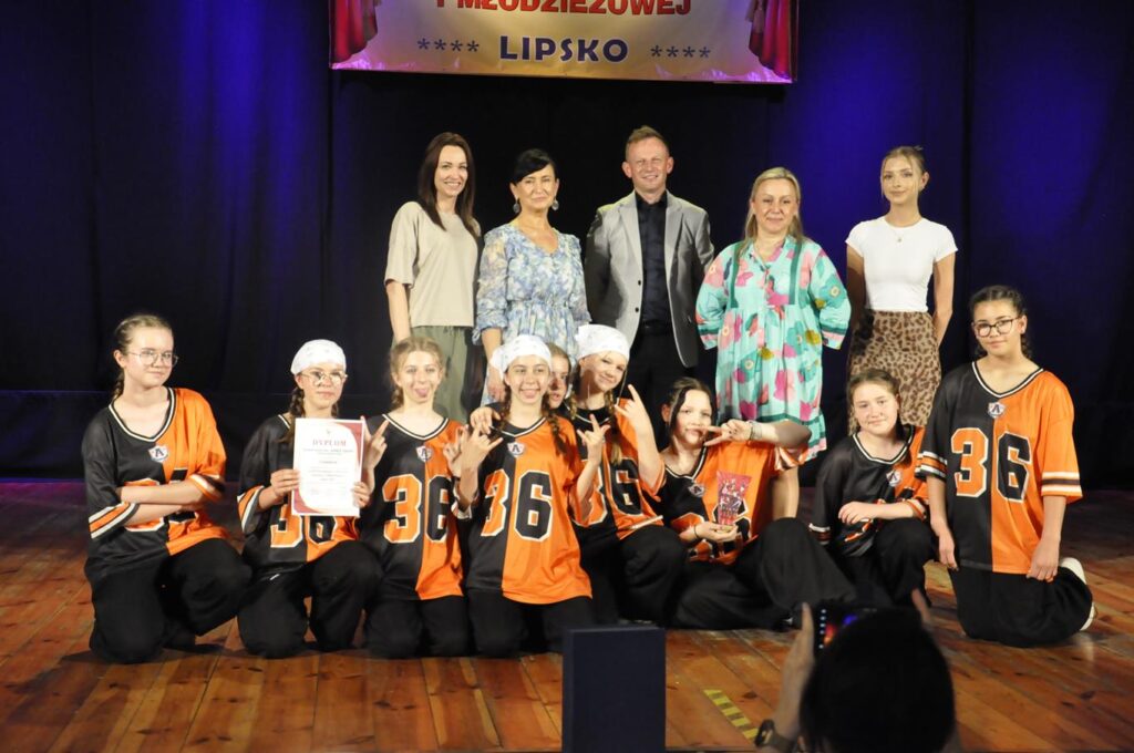 Grupa młodzieży w czarno-pomarańczowych koszulkach z numerami pozuje do wspólnego zdjęcia z kilkoma dorosłymi. Jedna osoba wznosi dyplom inna statuetkę. Niektórzy stoją inni siedzą. Nad nimi fragment baneru z nazwą wydarzenia. Tło czarno-granatowe.