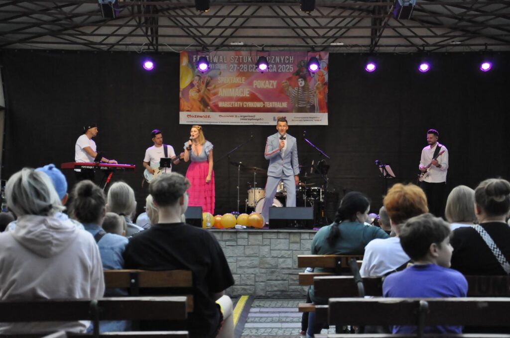 Zespół muzyczny w kolorowych strojach występuje na scenie plenerowej. Nad nimi wisi baner promujący wydarzenie, przed nimi siedzi publiczność na ławkach.