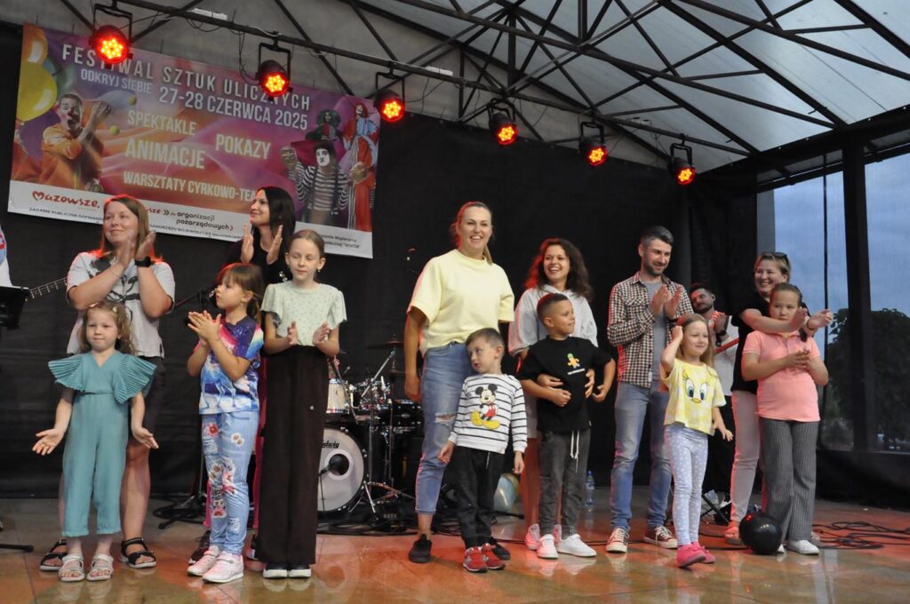 Grupa roześmianych dorosłych i dzieci stoi na scenie plenerowej. Niektórzy klaszczą. Za nimi instrumenty muzyczne oraz baner promujący wydarzenie.