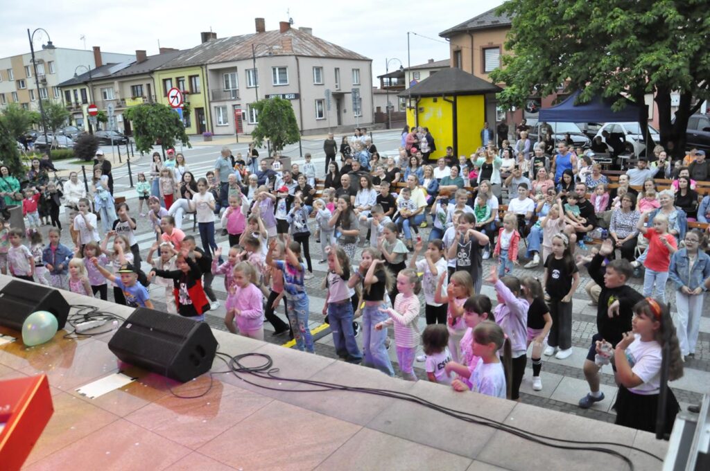 Duża grupa dzieci tańczy przed sceną plenerową. W tle fasady okolicznych budynków.