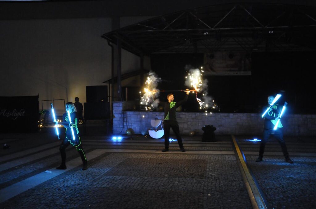 Troje artystów ulicznych w czarnych strojach z luminescencyjnymi seledynowymi wzorami trzyma zaświecone światła ledowe i urządzenia pirotechniczne. Tło w mroku.