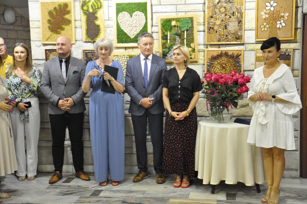 Kilkoro dorosłych w oficjalnych ubraniach stoi przy stole na którym stoi wazon z różowymi różami. Za nimi ściana z kolorowymi obrazami. Pomieszczenie ciepło oświetlone.