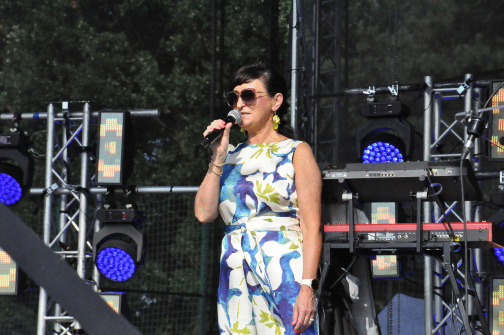 Kobieta w biało niebieskiej sukience i w okularach przeciwsłonecznych trzyma mikrofon przy ustach. W tle sprzęty elektroniczne i instrumenty muzyczne.