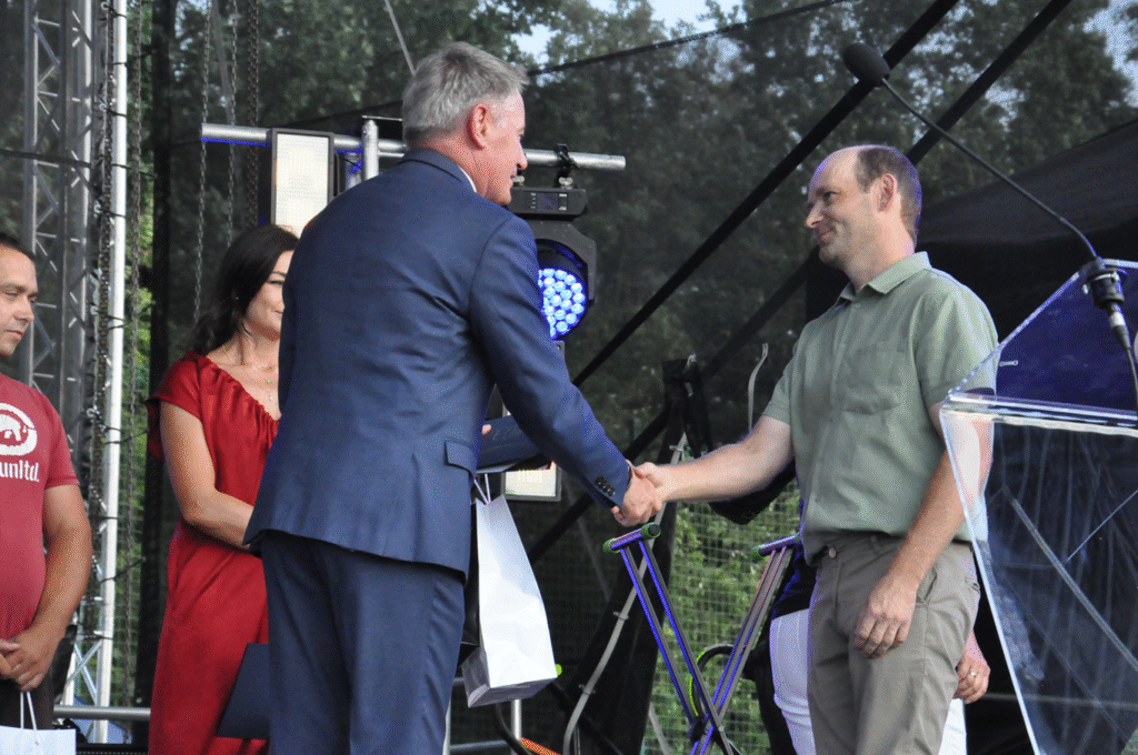 Mężczyzna w grafitowym garniturze podaje dłoń mężczyźnie w jasnozielonej koszuli i jasnych spodniach. Obok stoi kobieta w czerwonej sukience i mężczyzna w czerwonej koszulce. Za nimi sprzęty elektroniczne.