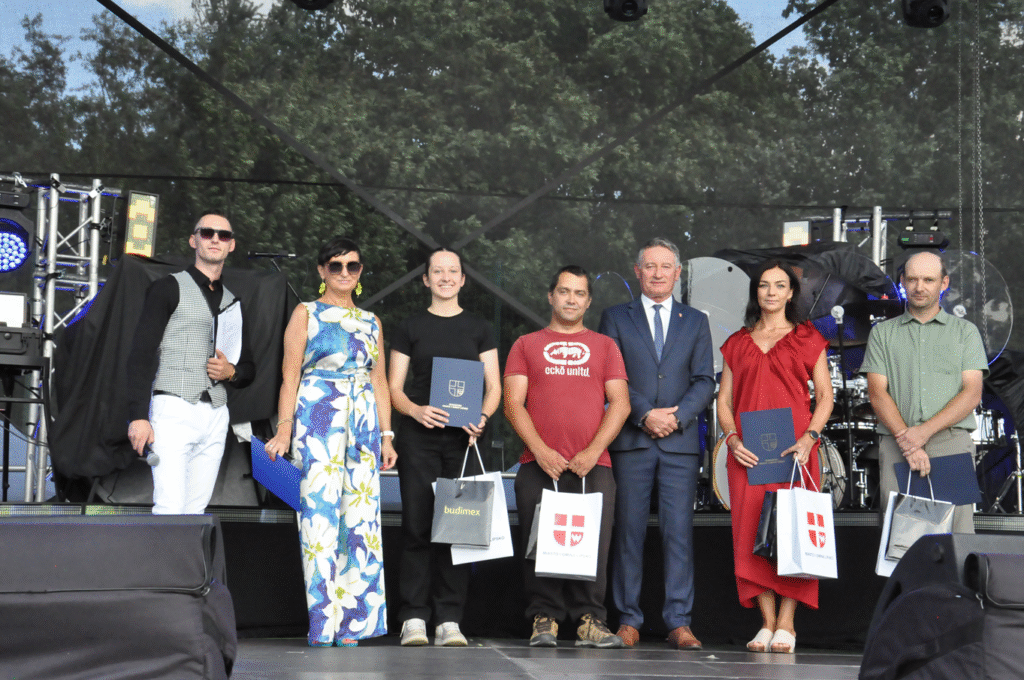 Grupa osób pozuje do wspólnego zdjęcia. Wszyscy stoją. Niektórzy trzymają torby z herbem miasta i logotypami