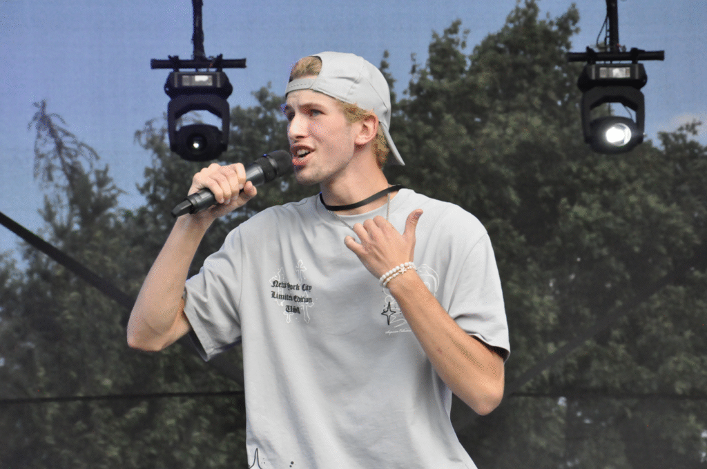 Chłopak w szarej koszulce i szarej czapce z daszkiem trzyma mikrofon przy ustach. Za nim korony drzew.