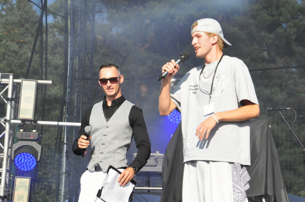 Chłopak w szarej koszulce i szarej czapce z daszkiem trzyma mikrofon przy ustach. Obok niego stoi mężczyzna w czarnej koszuli i kraciastej kamizelce, też trzyma mikrofon. Za nimi sprzęty elektroniczne.