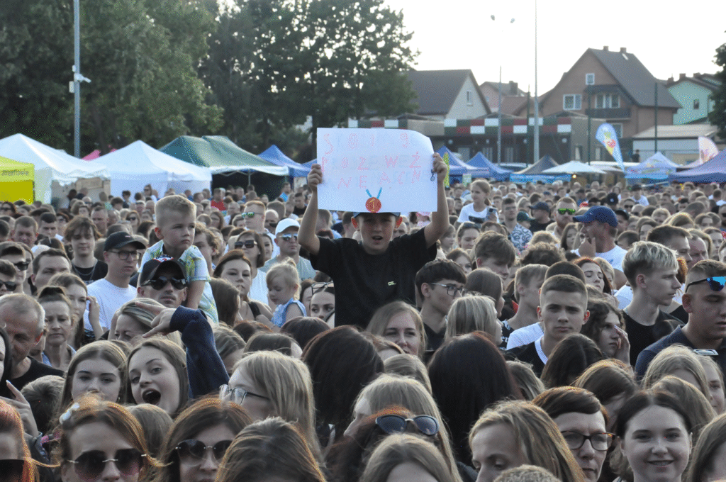 Tłum ludzi. W centrum zdjęcia chłopiec trzymający kartkę z napisem. Pogodny dzień.