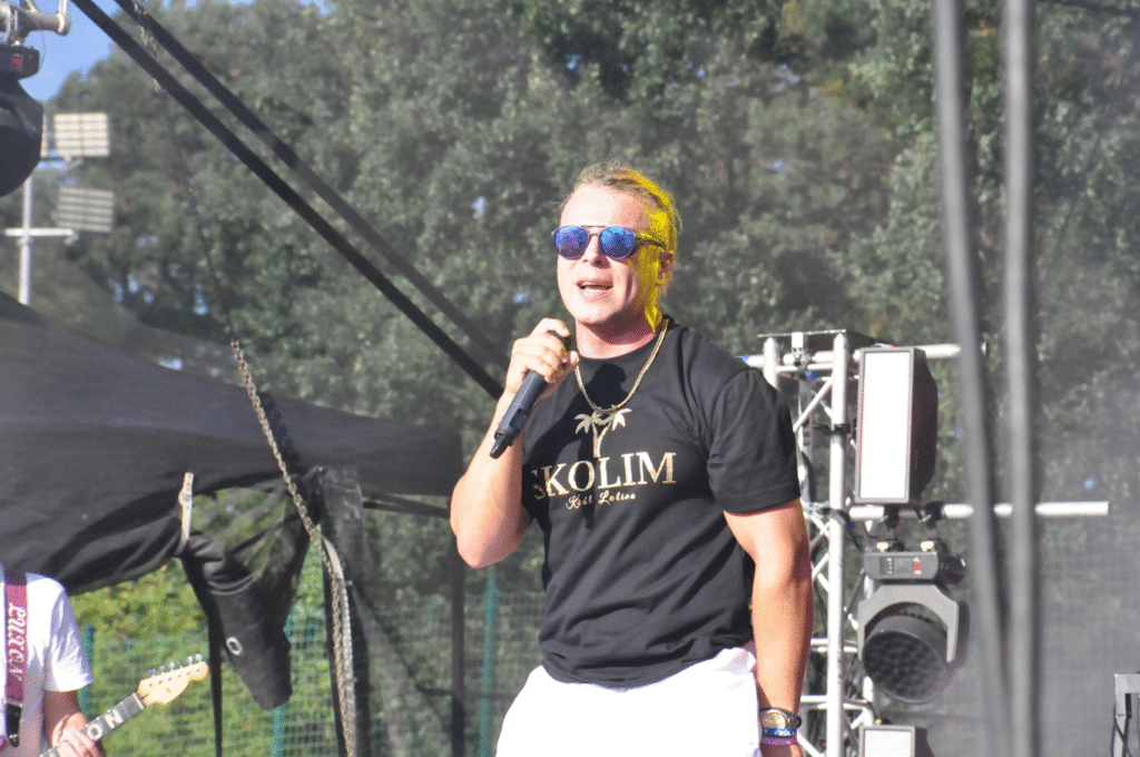 Mężczyzna w czarnej koszulce z napisem i okularach przeciwsłonecznych trzyma mikrofon przy ustach. Za nim sprzęty elektroniczne.