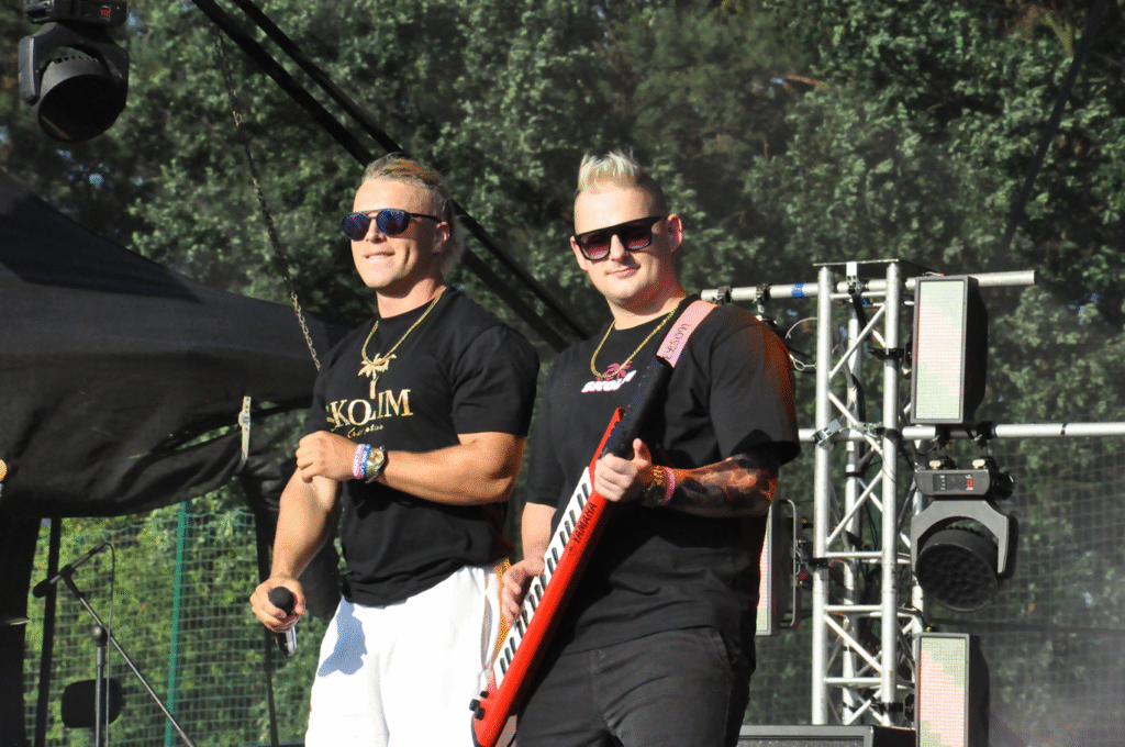 Mężczyzna w czarnej koszulce z napisem i okularach przeciwsłonecznych trzyma mikrofon. Obok niego mężczyzna w czarnej koszulce trzyma keyboard ręczny. Za nimi sprzęty elektroniczne.