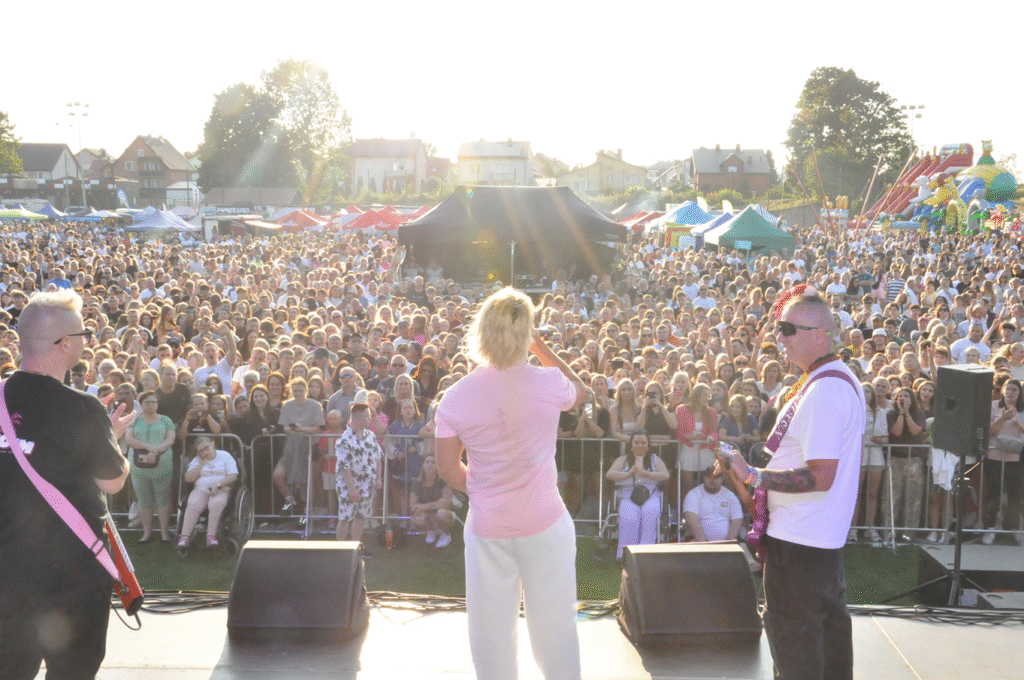 Na pierwszym planie artyści zwróceni w stronę tłumnie zgromadzonej publiczności. Słoneczny dzień.