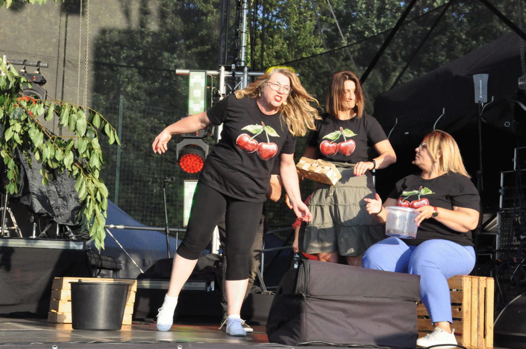 Trzy kobiety z koszulkami z nadrukami wiśni. Jedna siedzi, dwie stoją. Trzymają koszyki albo wiaderka. Słoneczny dzień.