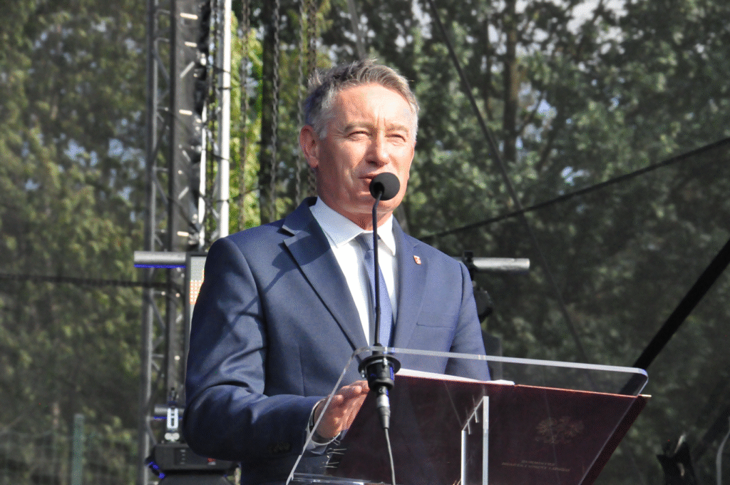 Mężczyzna w grafitowej marynarce stoi przy mównicy z mikrofonem na statywie. W tle korony drzew.