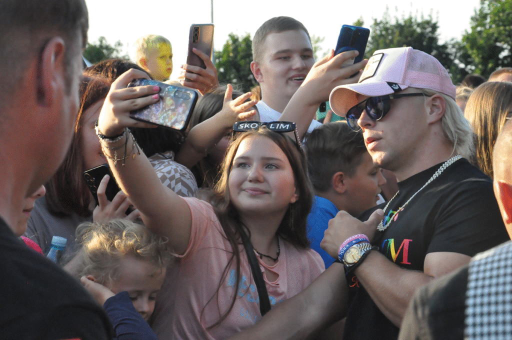 Mężczyzna w okularach przeciwsłonecznych i różowej czapce z daszkiem pozuje do wspólnego zdjęcia z fanką.. Obok stoi wiele innych roześmianych osób.