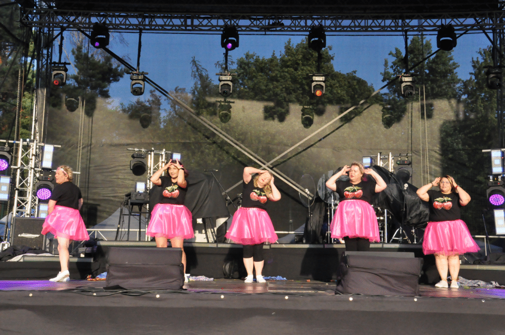 Pięć kobiet w czarnych koszulkach z nadrukiem wiśni i różowych sukienkach tańczy na scenie.