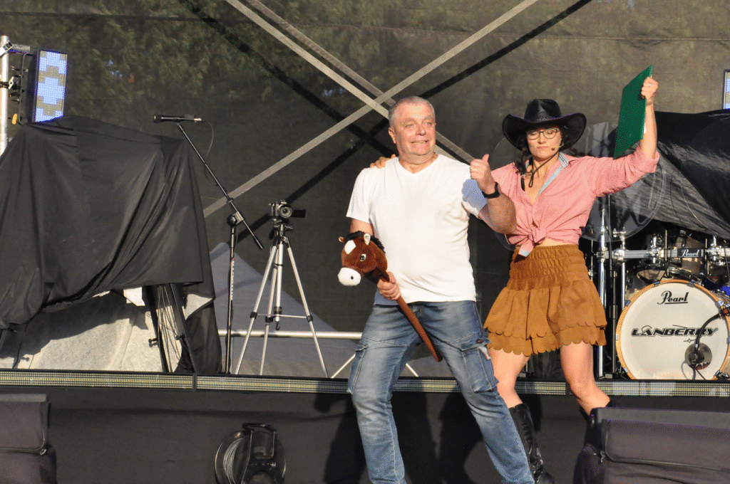 Mężczyzna z kijkiem z głową konia i cowbojka tańczą na scenie. Za nimi sprzęty elektroniczne i instrumenty muzyczne.