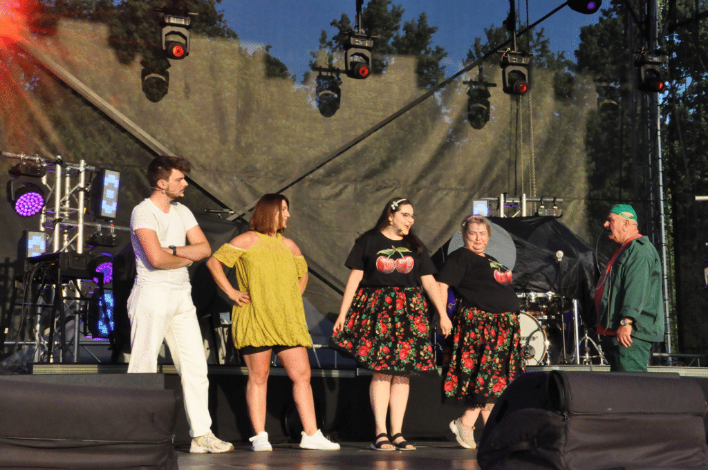 Kila osób w kolorowych strojach przypominających owoce stoi na scenie. Za nimi sprzęty elektroniczne i instrumenty muzyczne.