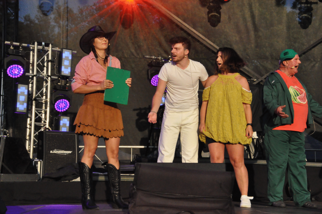 Cztery osoby w kolorowych strojach. Jedna osoba ma strój cowbojki. Za nimi sprzęty elektroniczne i instrumenty muzyczne.