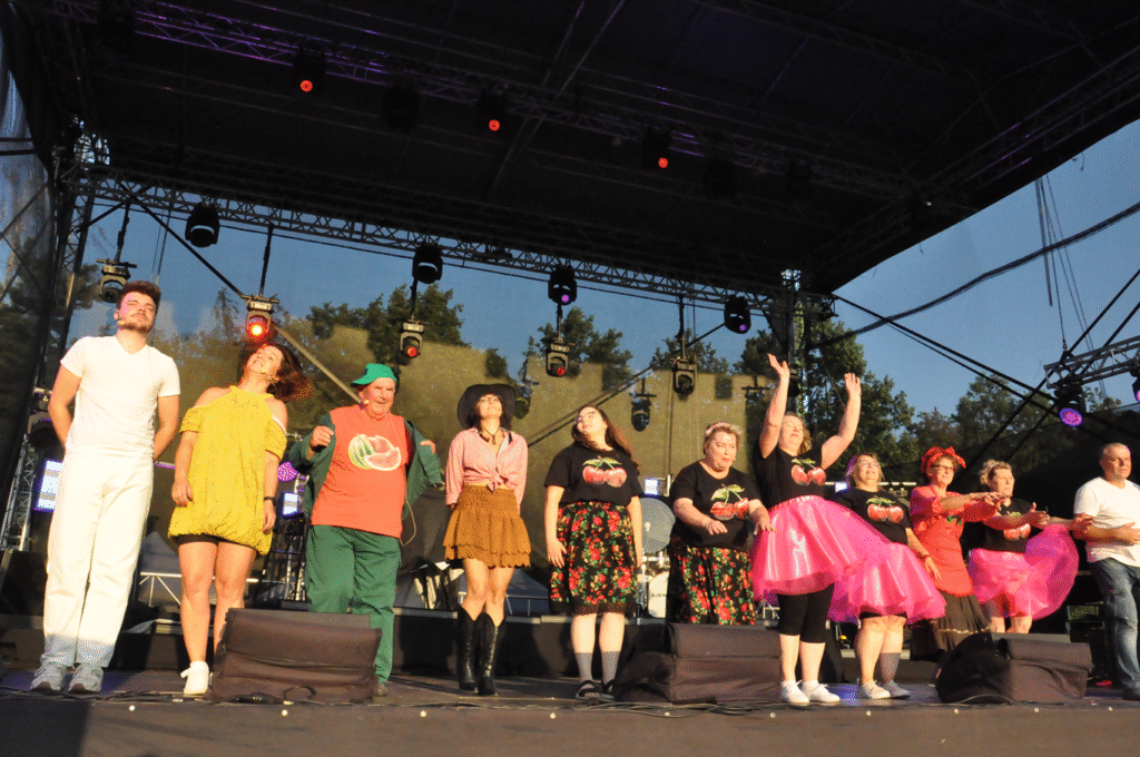 Duża grupa osób w kolorowych strojach tańczy na scenie. Za nimi sprzęty elektroniczne.