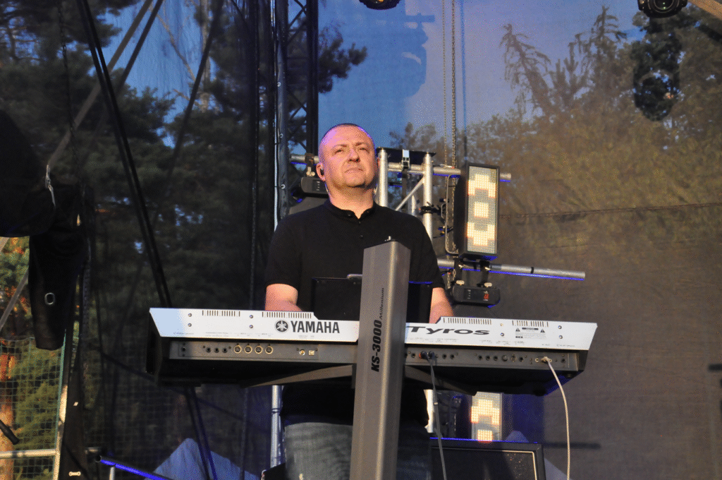 Mężczyzna w czarnej koszulce stoi przy keyboardzie.. Za nim fragmenty sceny.