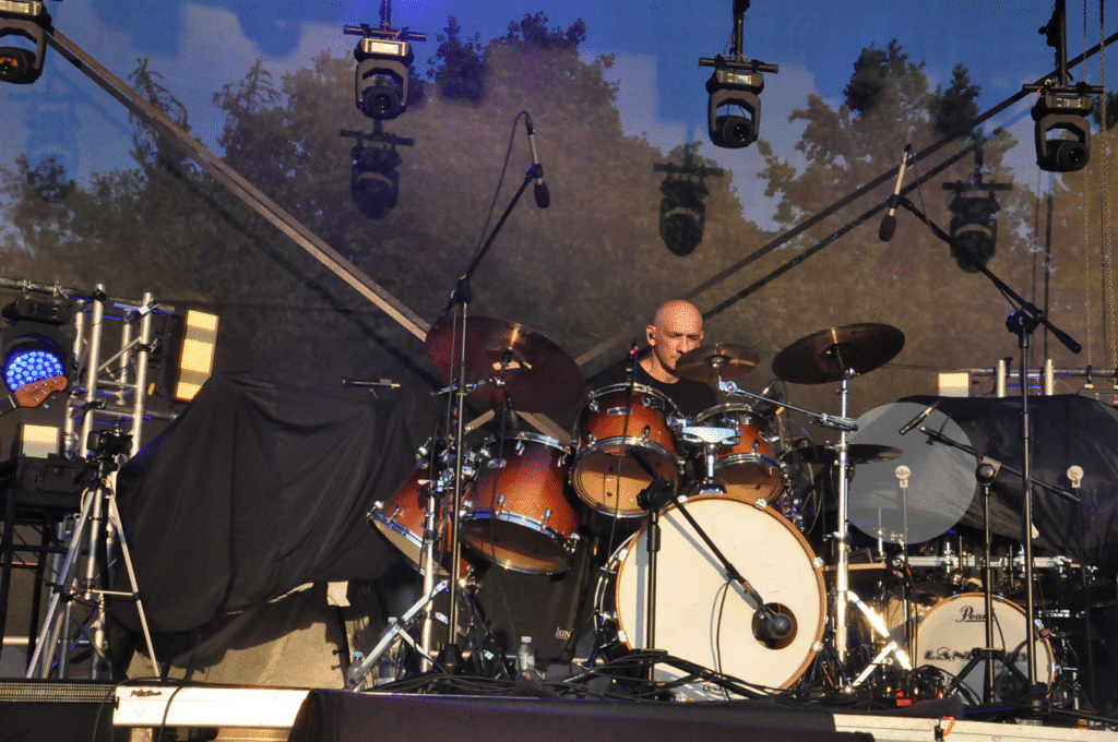 Łysy mężczyzna w czarnej koszulce gra na perkusji na scenie.