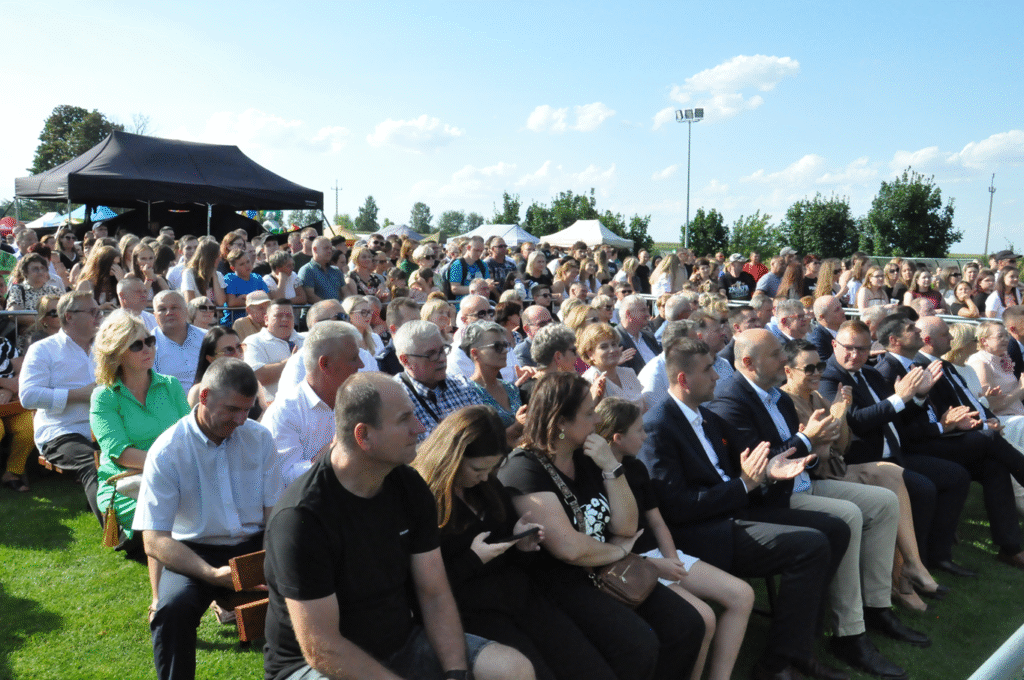 Tłum ludzi. Niektórzy siedzą, niektórzy siedzą na łakach. W tle namioty z atrakcjami wydarzenia.