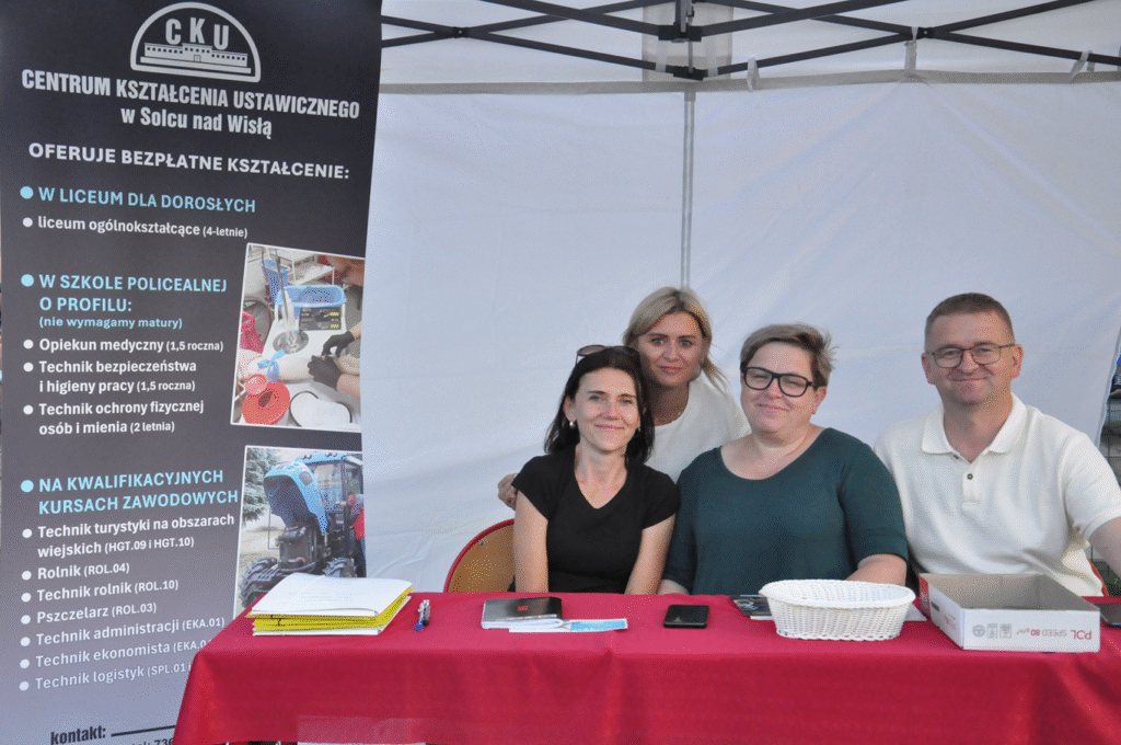 Trzy kobiety i mężczyzna siedzą przy stole okrytym czerwonym obrusem, na którym leża dokumenty, długopisy, koszy. bok nich stoi rollup z reklamą szkoły.