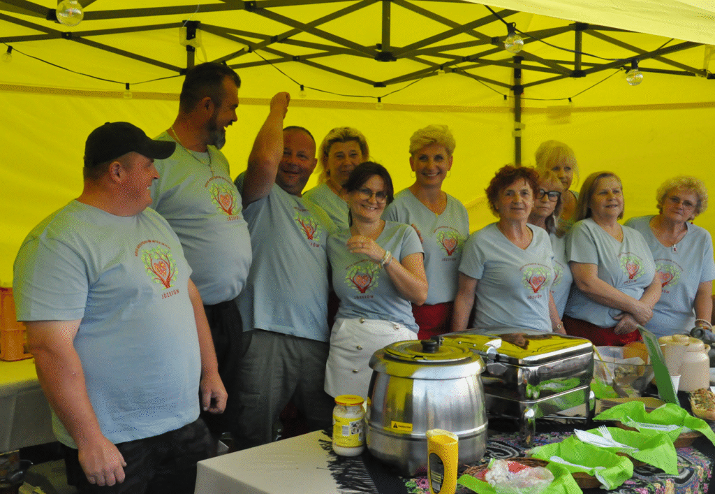 Duża grupa osób, kobiety i mężczyźni w szarych koszulkach z logotypem KGW pozuje do wspólnego zdjęcia pod żółtym namiotem. Przed nimi asortyment kuchni regionalnej..