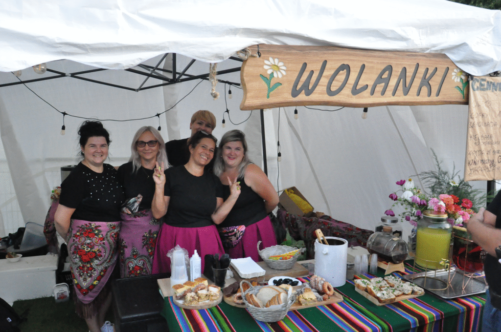 Kobiety w czarnych koszulkach i różowych spódniczkach pozują do wspólnego zdjęcia. Przed nimi asortyment kuchni regionalnej na kolorowym kocu.