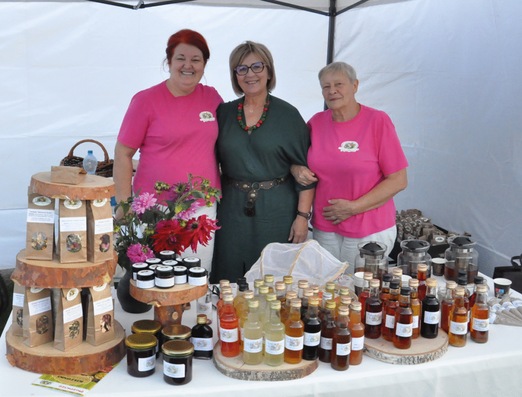 Trzy kobiety, dwie w różowych koszulkach , jedna w szarej sukience pozują do wspólnego zdjęcia. Przed nimi stoją słoiczki i butelki z wyrobami kuchni regionalnej.