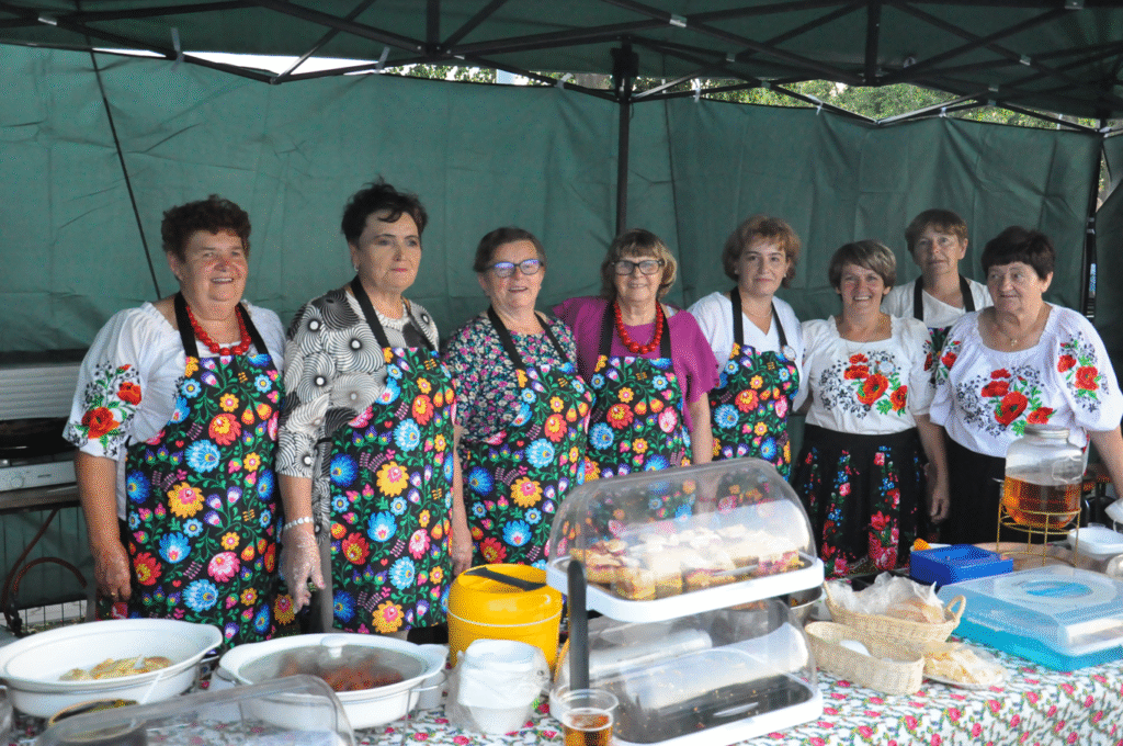 Kilka kobiet w kolorowych fartuszkach z motywem folkloru pozuje do wspólnego zdjęcia pod zielonym namiotem. Przed nimi wyroby kuchni regionalnej.
