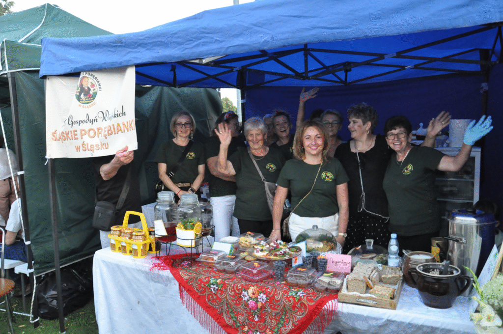 Kobiety w zielonych koszulkach z logotypem KGW pozują do wspólnego zdjęcia. Przed nimi na stole okrytym czerwoną chustą leżą wyroby kuchni regionalnej. Obok nich wisi mały baner z nazwą KGW.