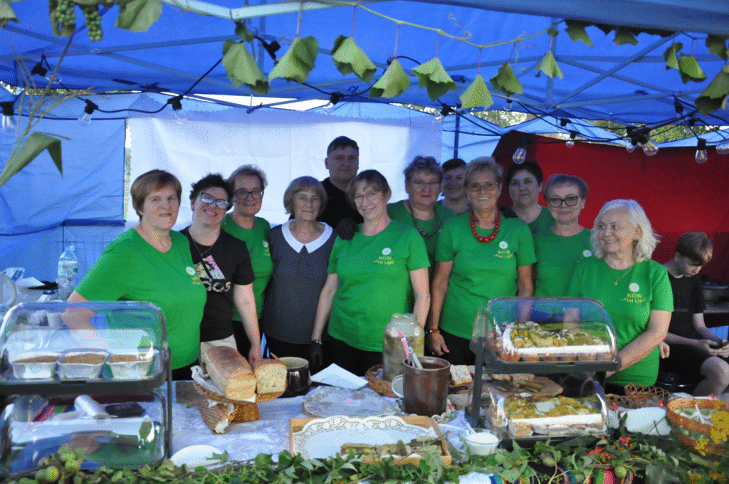 Duża grupa kobiet w zielonych koszulkach i jeden mężczyzna z uśmiechami na twarzach pozują do wspólnego zdjęcia. Przed nimi na stole okrytym białym obrusem i zieleniną leżą wyroby kuchni regionalnej.