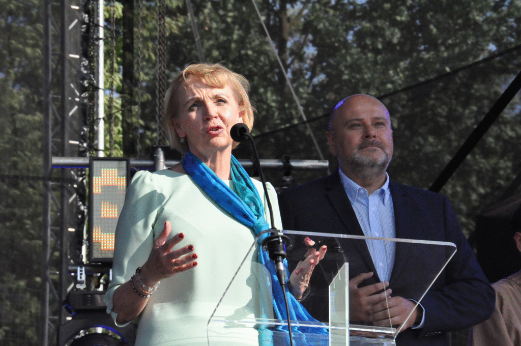 Kobieta w jasnozielonej sukience i niebieskiej apaszce mówi do mikrofonu na statywie. Za nią stoi mężczyzna w grafitowej marynarce. W tle korony drzew.