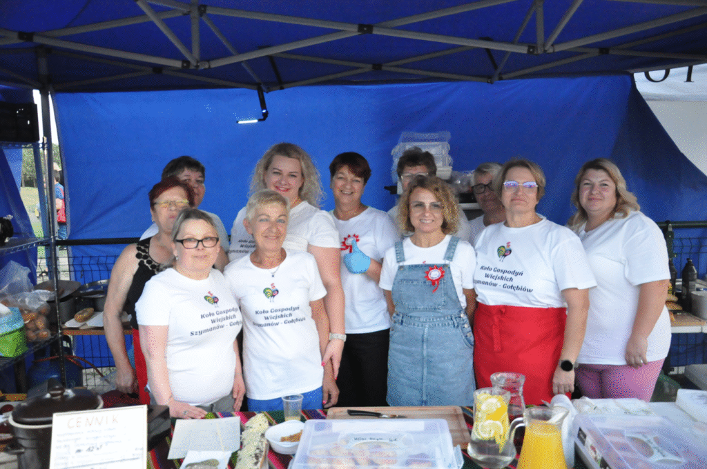 Kobiety w białych koszulkach z nazwą swojego KGW z uśmiechami pozują do wspólnego zdjęcia pod niebieskim namiotem. Przed nimi leżą wyroby kuchni regionalnej.
