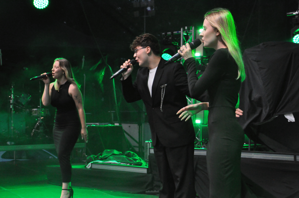 Dwie młode kobiety i młody mężczyzna w czarnych strojach trzymają mikrofony przy ustach. Za nimi sprzęty elektroniczne. Scena w półmroku.