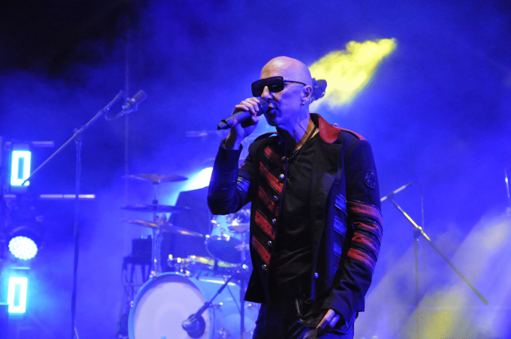 Łysy mężczyzna w okularach przeciwsłonecznych i czarno-czerwonej estradowej kurtce trzyma mikrofon przy ustach. Scena oświetlona jaskrawym światłem.