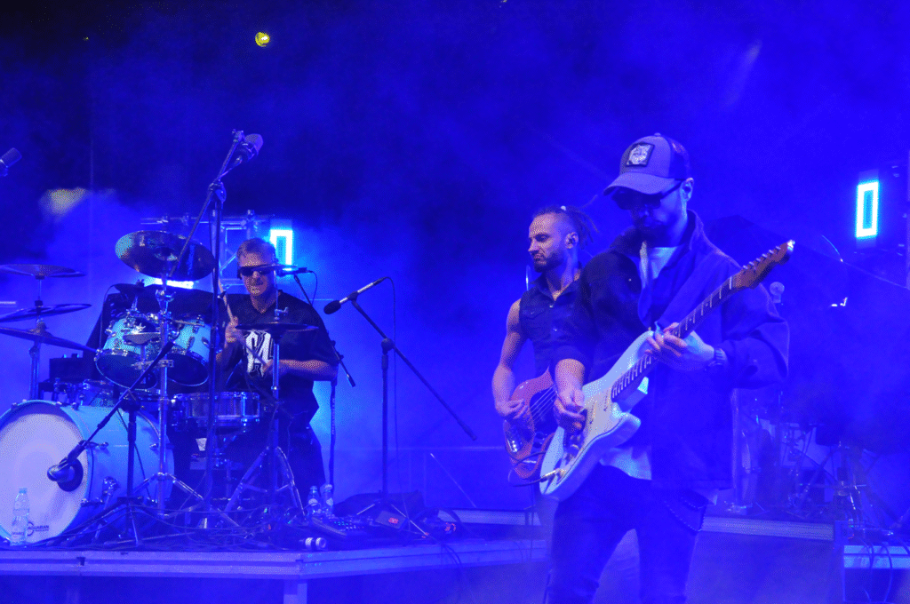 Troje muzyków. Jeden gra na perkusji, dwóch na gitarach elektrycznych. Scena oświelona jaskrawym światłem.