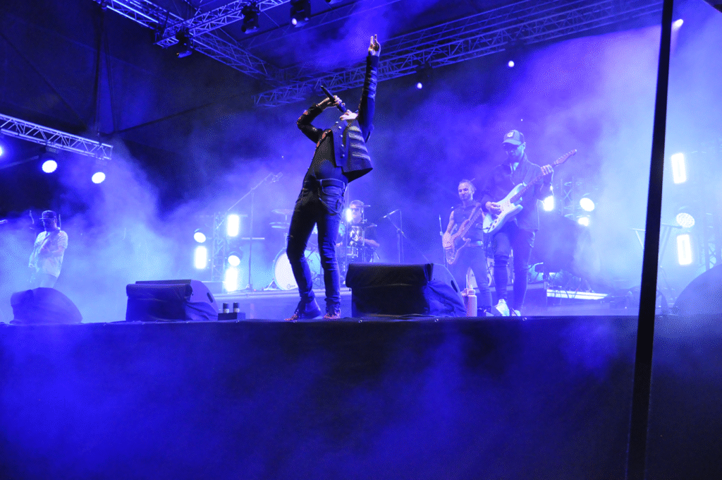 4 muzyków na scenie. Frontman wznosi ręce do góry. Wieczorna scena plenerowa.