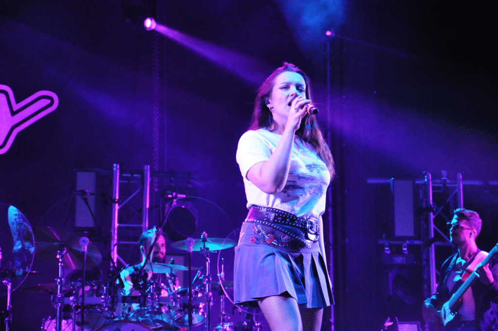 Kobieta w jasnej koszulce i krótkiej szarej sukience trzyma mikrofon przy ustach. Za nią członkowie zespołu grają na instrumentach. Wieczorna scena.